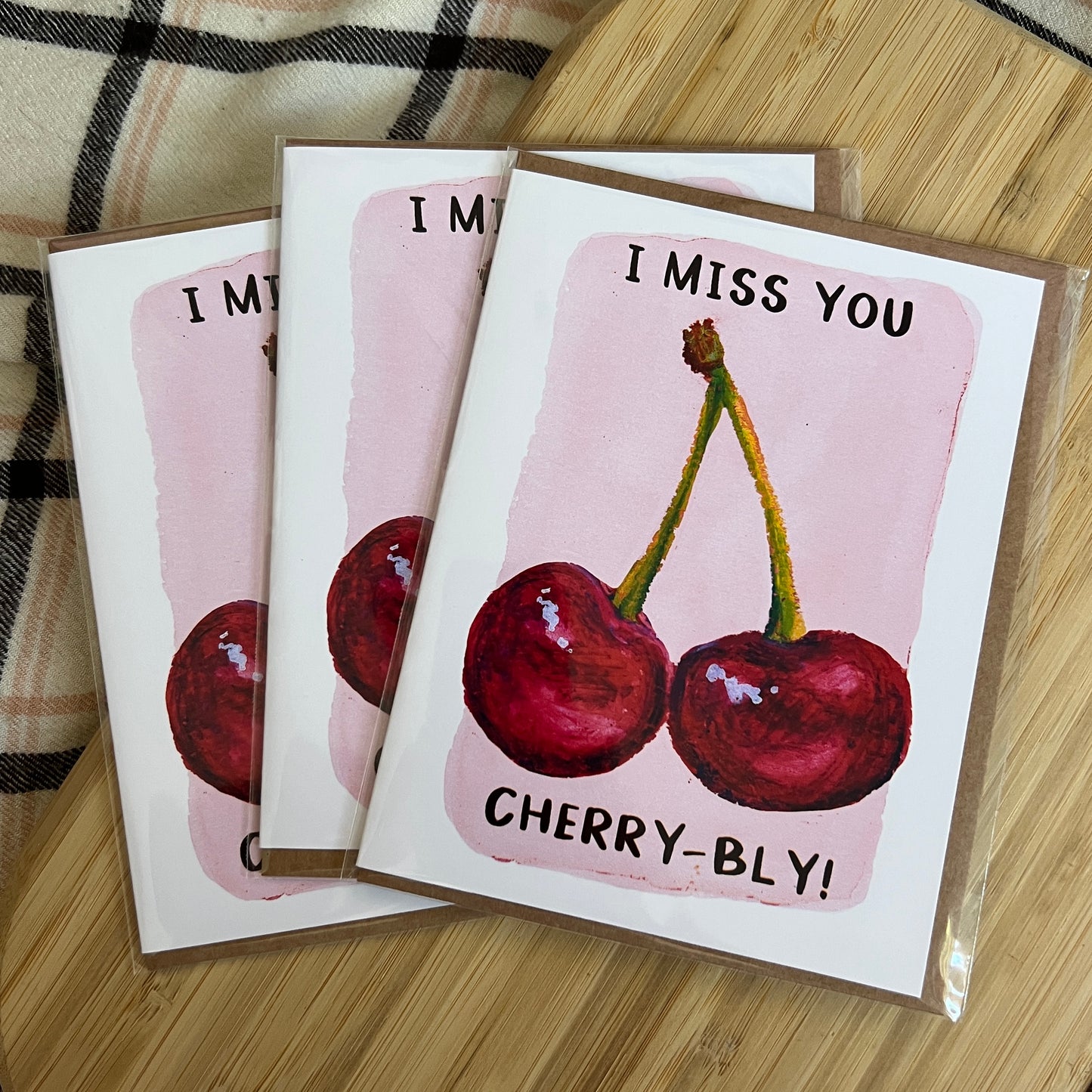 Three 'I Miss You Cherry-bly!' greeting cards on a wooden surface with a plaid fabric background.