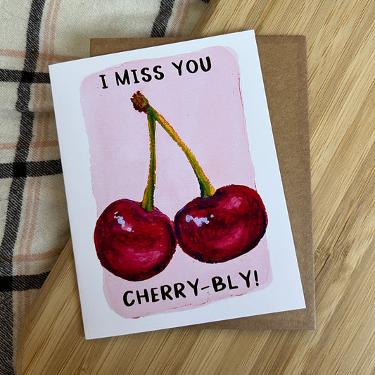 Card with cherries and text 'I Miss You Cherry-bly!' on a wooden surface.