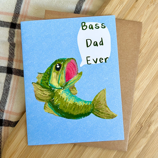 Card with a fish illustration and 'Bass Dad Ever' text on a wooden surface.