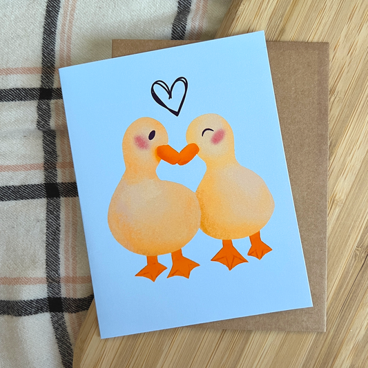 Card with two cartoon ducks holding hands and a heart above them on a light blue background.