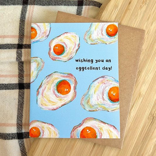 Greeting card with omelette design and text 'wishing you an eggcellent day!' on a wooden surface.