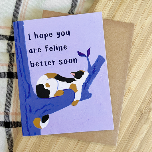 Greeting card with a cat illustration and text 'I hope you are feline better soon' on a wooden surface