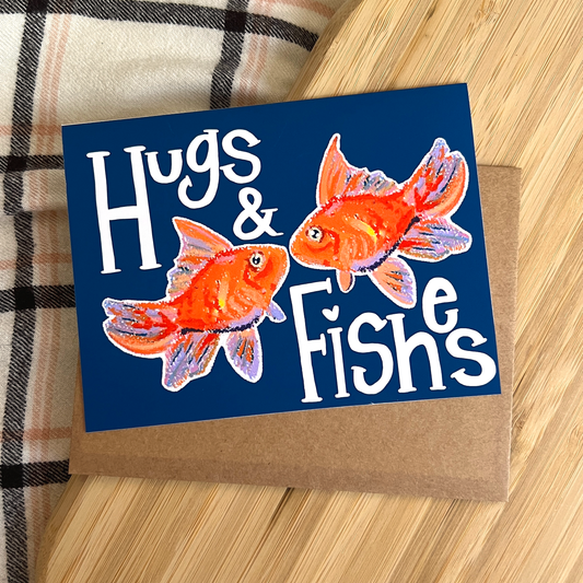Greeting card with goldfish illustration and text 'Hugs & Fishes' on a wooden surface.