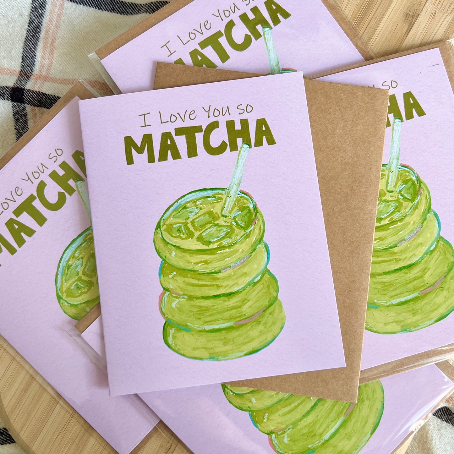 Greeting card with 'I Love You So Matcha' text and matcha illustration on a wooden surface.