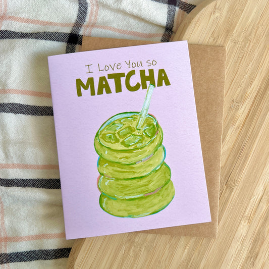 Card with matcha illustration and text 'I Love You So Matcha' on a wooden surface.