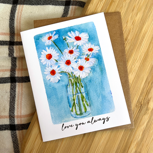 Greeting card with daisy design and 'love you always' text on a wooden surface.