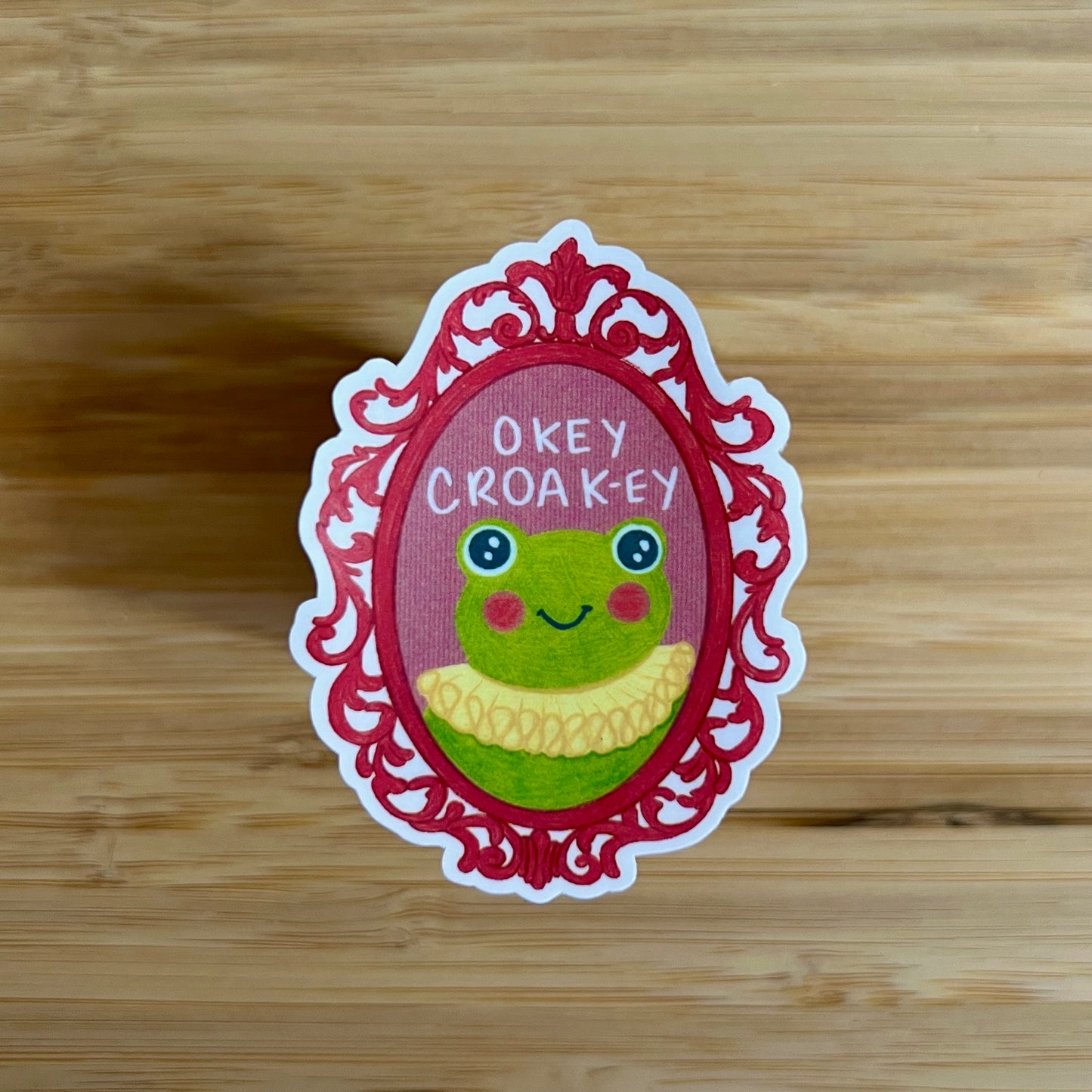 Sticker with a frog design and text 'Okey Croak-ey' on a wooden surface