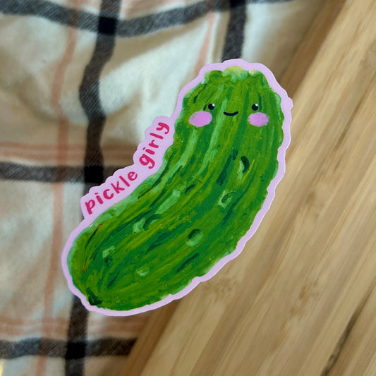 Sticker of a green pickle with facial features and 'pickle girly' text on a wooden surface.
