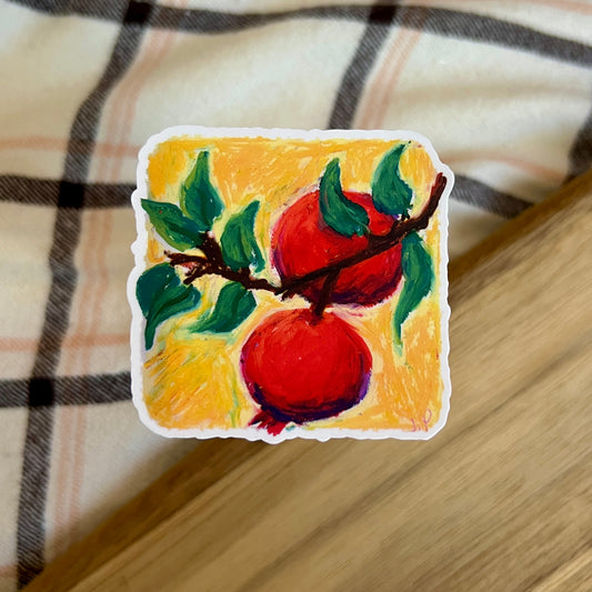 Sticker with a painting of two red pomegranates on a branch against a yellow background, placed on a checkered fabric surface.