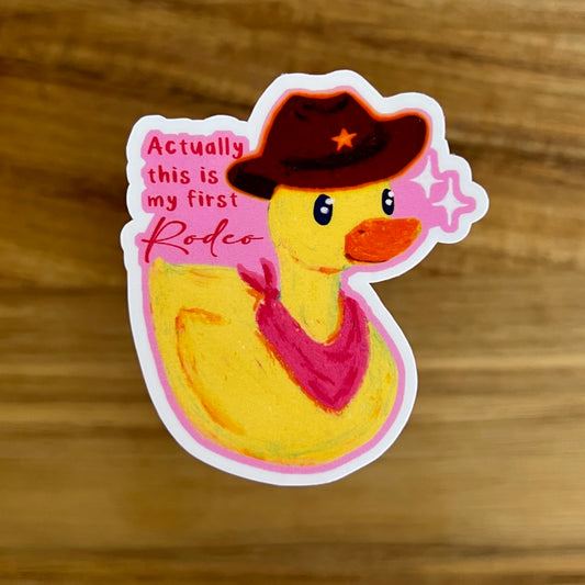Sticker of a duck wearing a cowboy hat and bandana with text 'actually this is my first rodeo' on a wooden surface