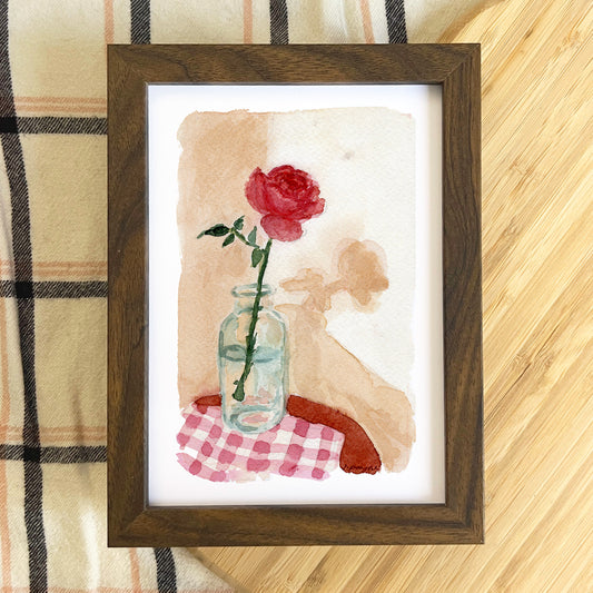 Framed artwork of a red rose in a glass bottle on a checkered cloth with a wooden frame.