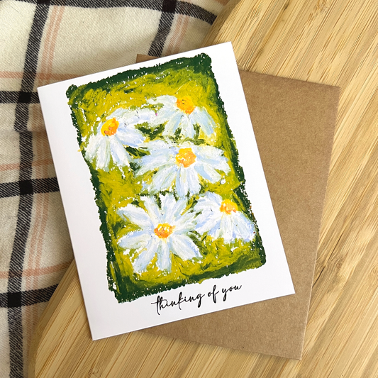 Greeting card with daisy design and 'thinking of you' text on a wooden surface.