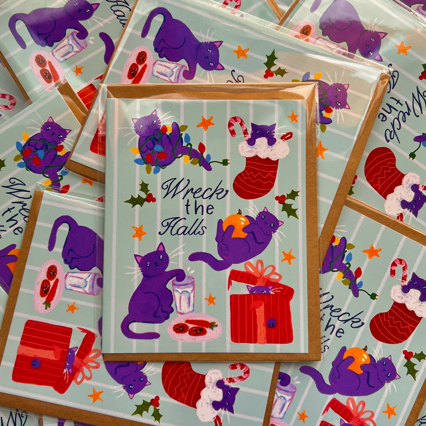 Stack of greeting cards with purple cats and Christmas-themed illustrations on a light blue background.