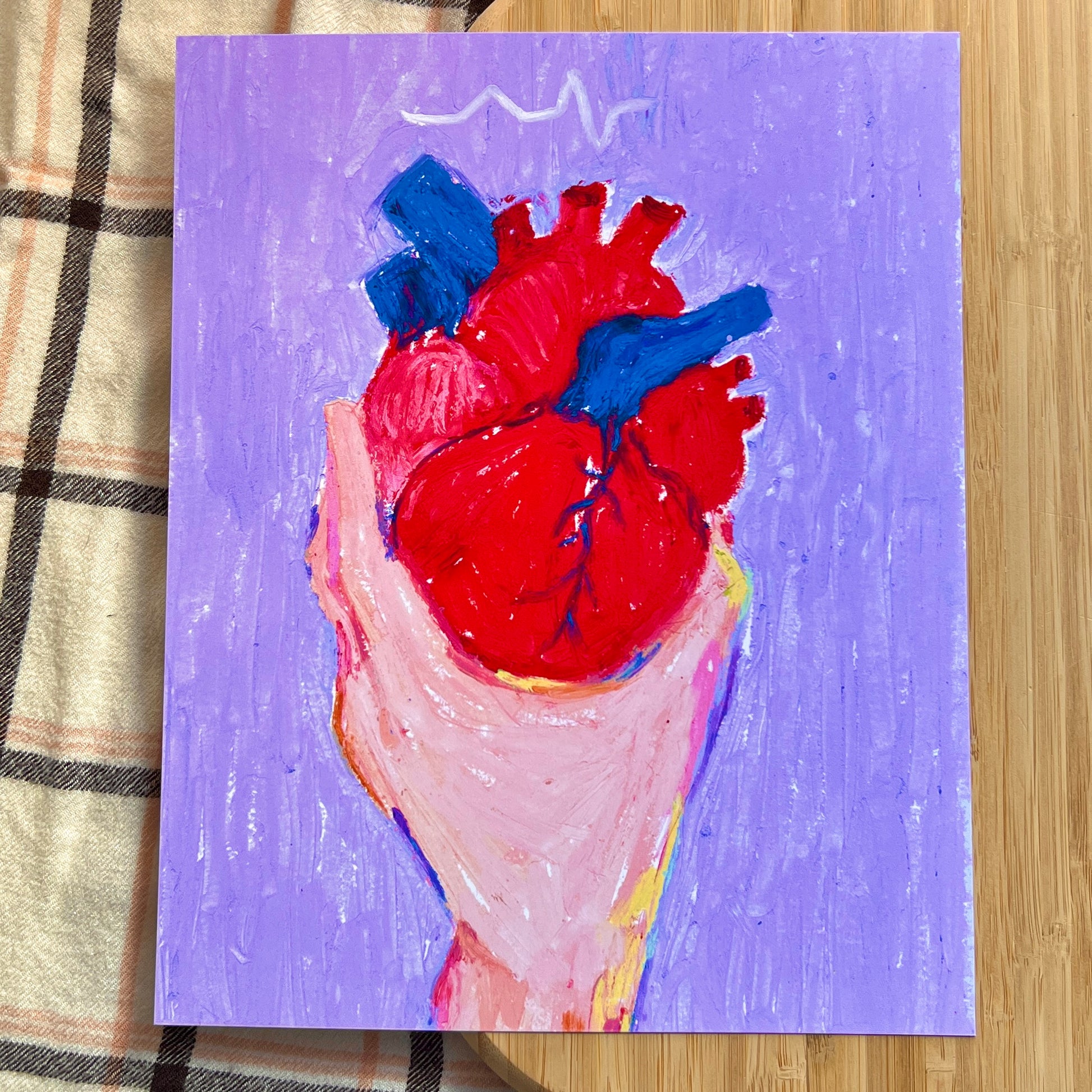 hand holding anatomical heart with purple background on a wooden and plaid surface