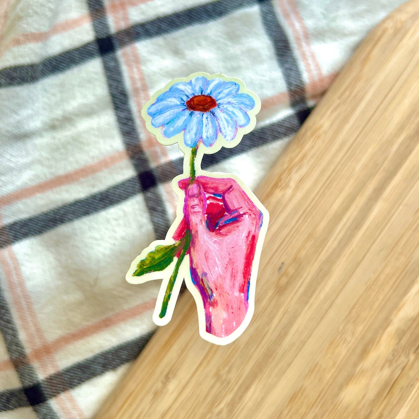 Sticker of a hand holding a daisy on a wooden surface.