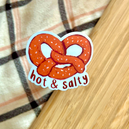 Sticker of a pretzel with 'hot & salty' text on a wooden surface.