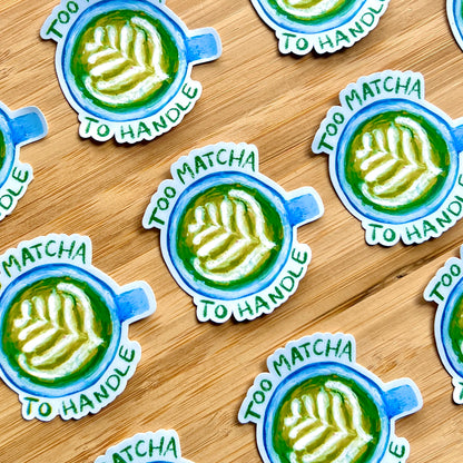 Stickers with a matcha latte design and text 'Too Matcha To Handle' on a wooden surface.