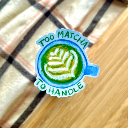Sticker with matcha design and text 'Too Matcha To Handle' on a wooden surface.