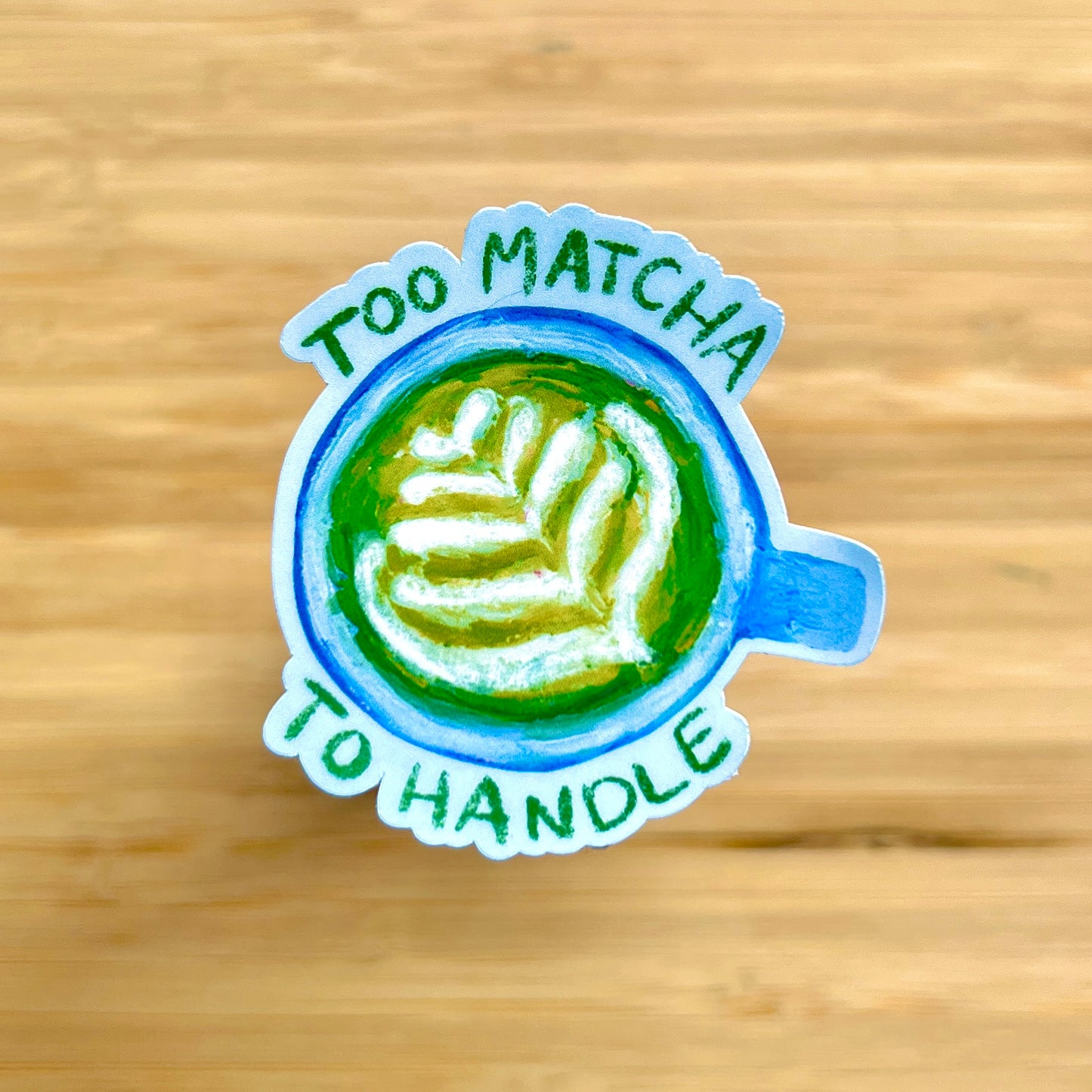 Sticker with a matcha latte design and text 'Too Matcha To Handle' on a wooden surface.