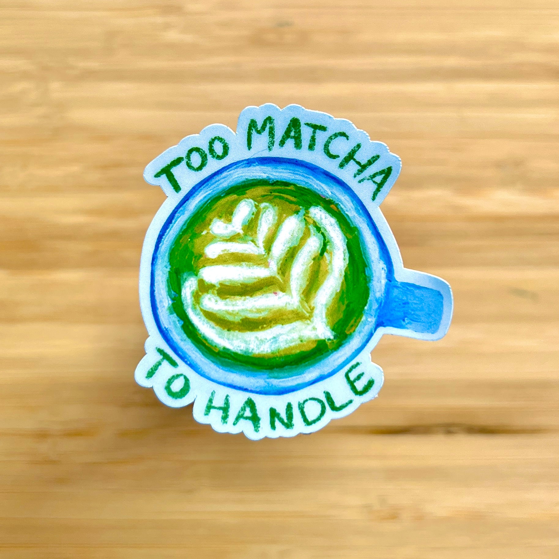 Sticker with a matcha latte design and text 'Too Matcha To Handle' on a wooden surface.
