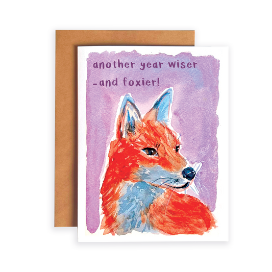 Greeting card with a fox illustration and text 'another year wiser and foxier!' on a purple background.