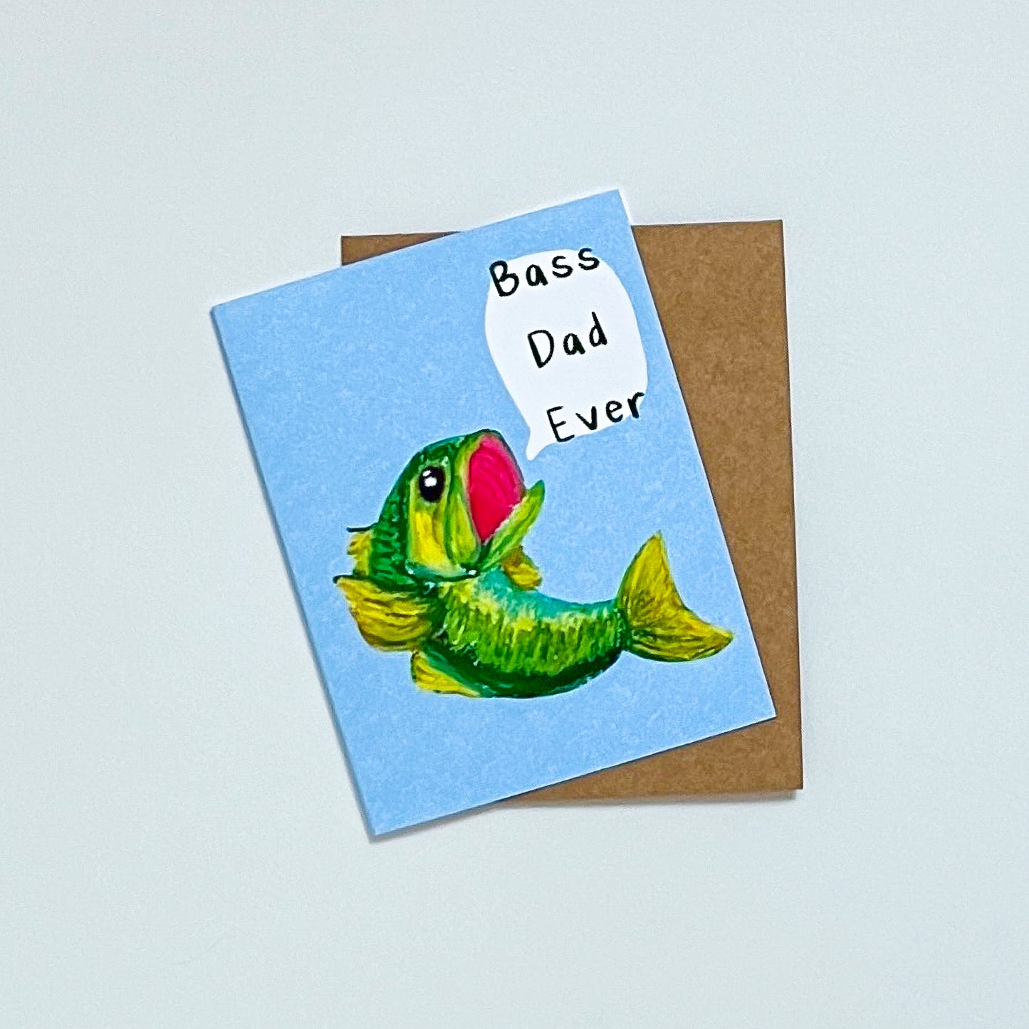 greeting card design of  a bass fish and text 'bass dad ever' on it. On top of a Kraft envelope with white background