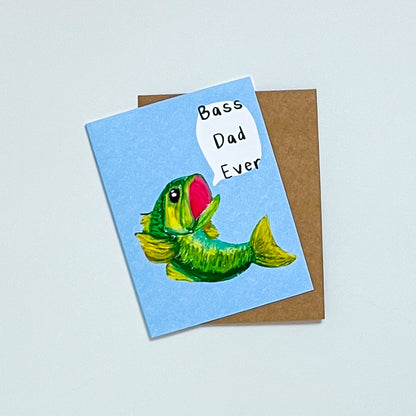 greeting card design of  a bass fish and text 'bass dad ever' on it. On top of a Kraft envelope with white background