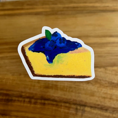 Sticker of a slice of pie with blueberry topping on a wooden surface