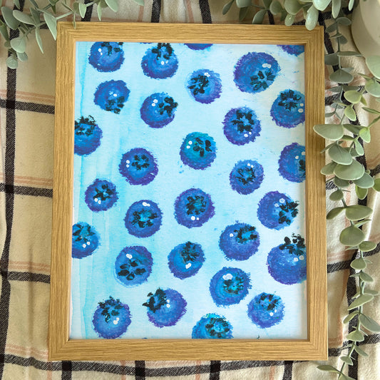 Framed artwork with blueberry pattern on a light blue background, placed on a checkered fabric surface.