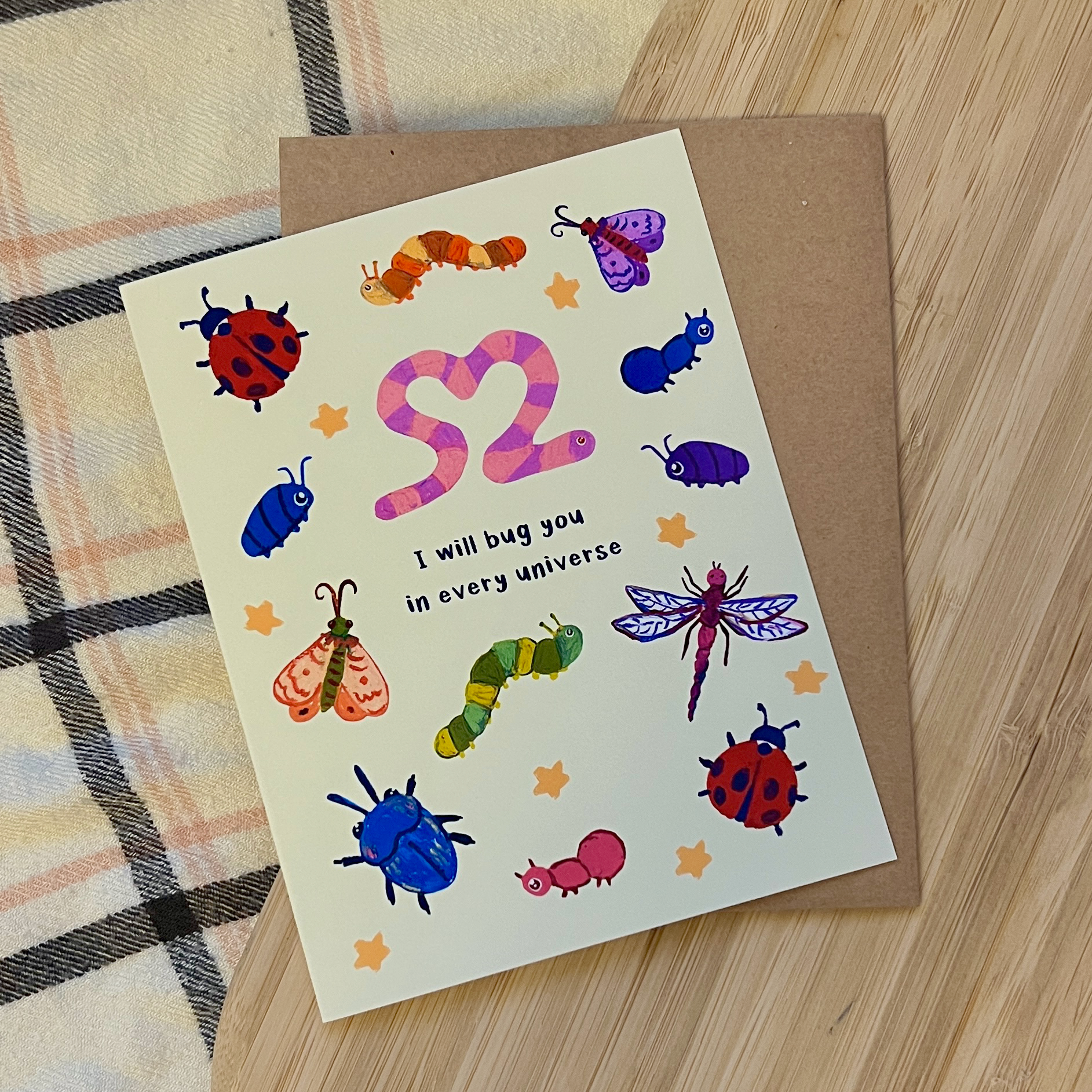 Greeting card with colorful insect illustrations and text on a wooden surface.