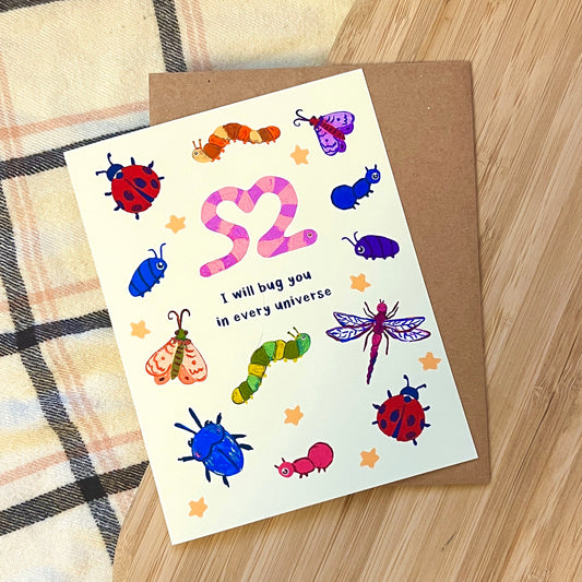 Greeting card with colorful insect illustrations and text 'I will bug you in every universe' on a wooden surface.