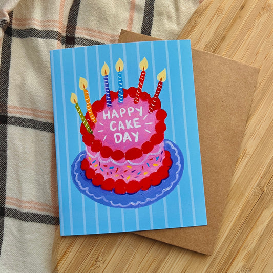 Birthday card with a cake illustration and 'Happy Cake Day' text on a blue background, placed on a wooden surface.