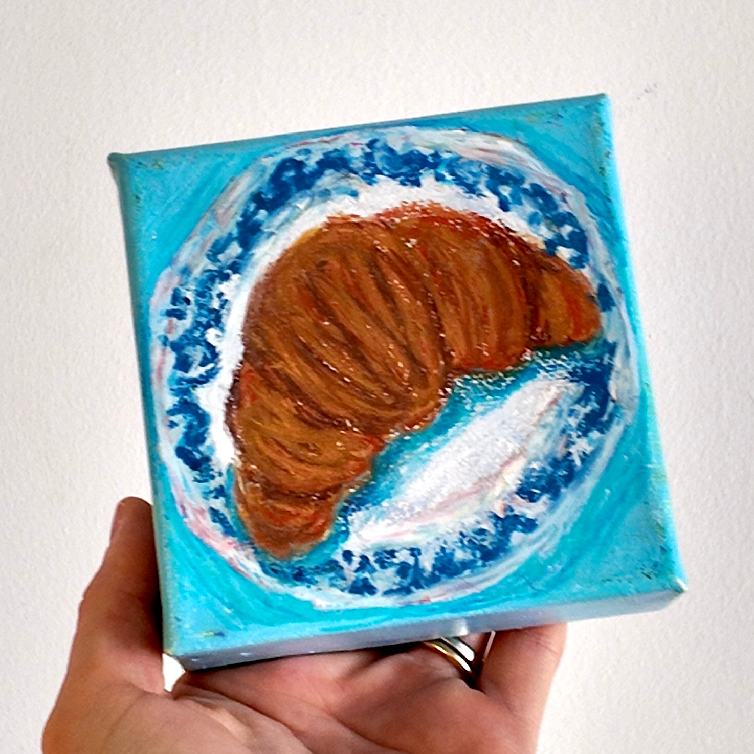 Hand holding a small square canvas with a croissant painting against a white background