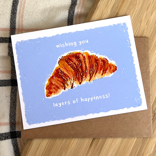 Greeting card with a croissant illustration and text on a wooden surface