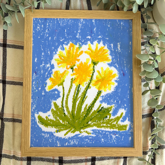 Framed artwork of yellow flowers on a blue background, leaning against a plaid fabric background.