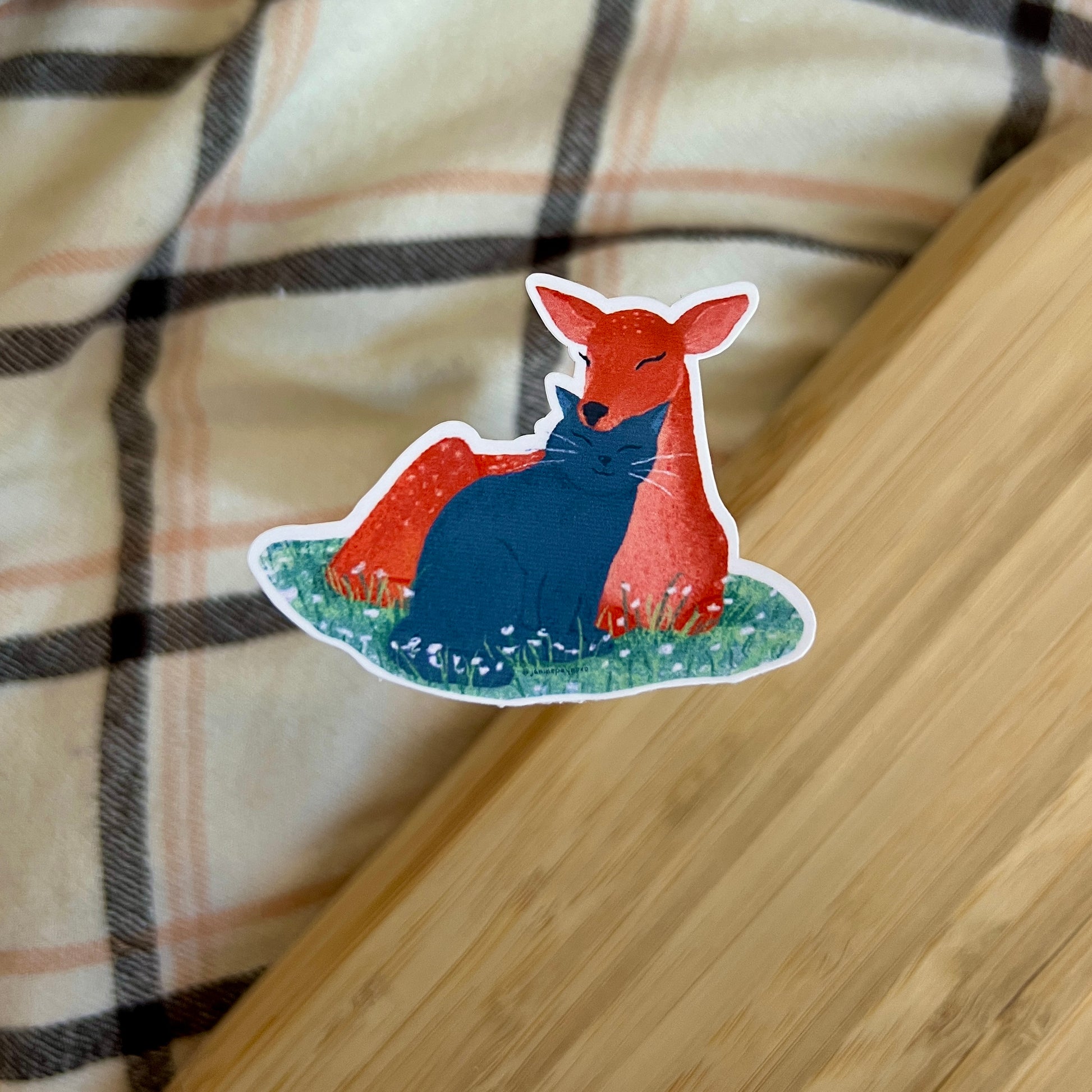 Sticker of a deer and cat on a wooden surface with plaid fabric in the background