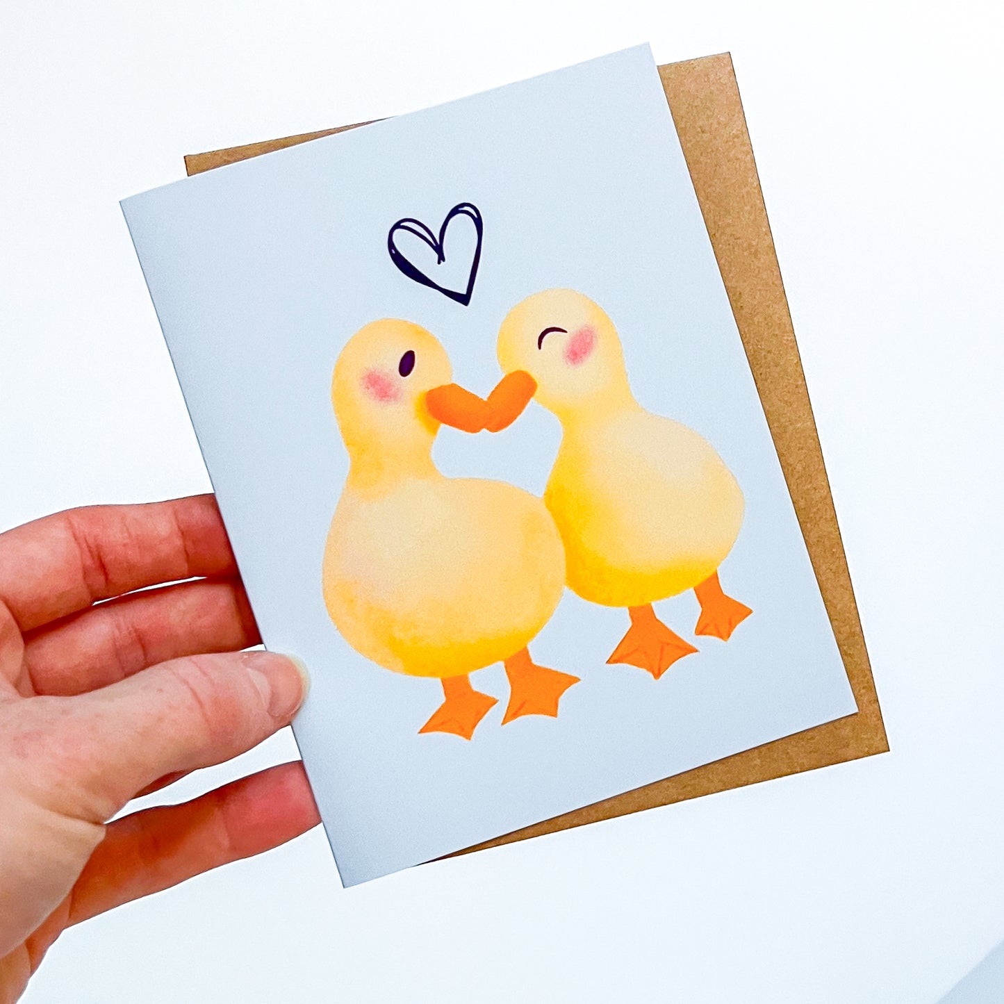 Greeting card with two ducks holding hands and a heart above them, held by a hand.
