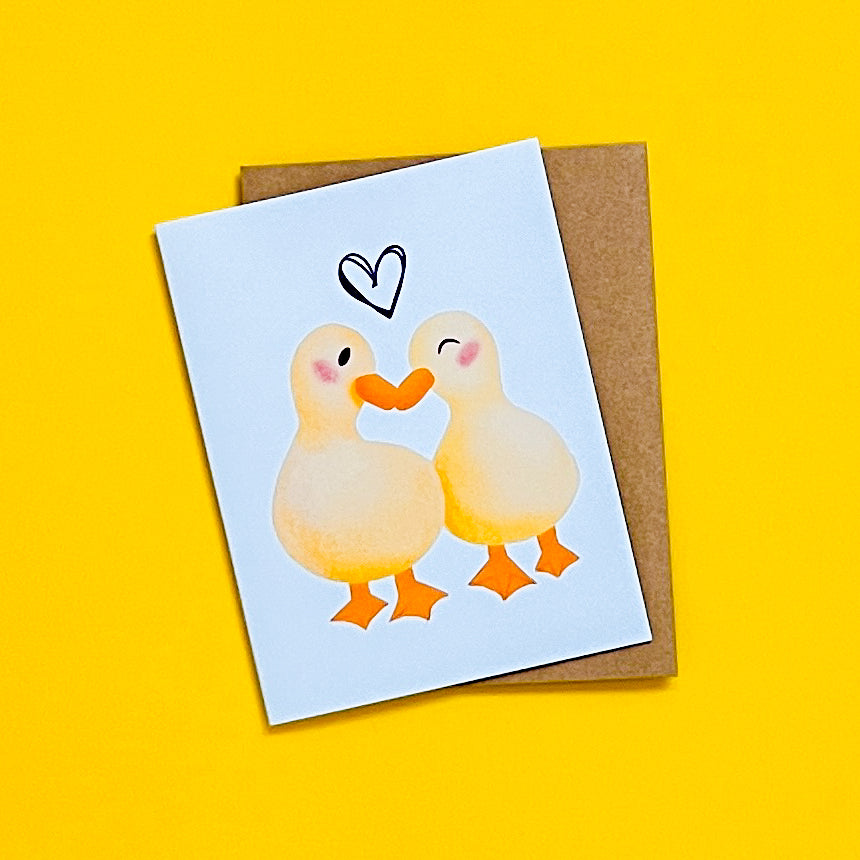 Greeting card with two ducks holding hands and a heart on a yellow background