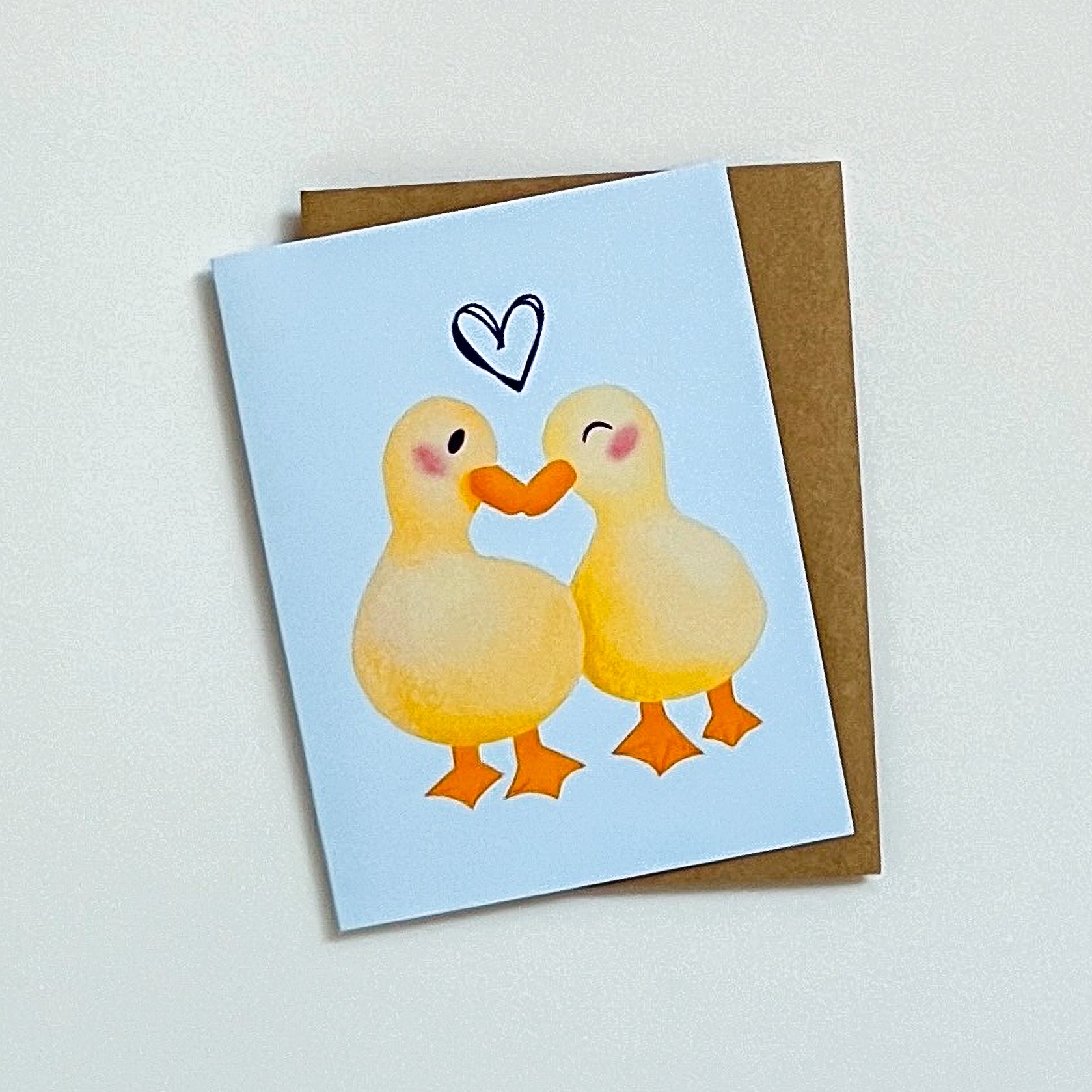Greeting card with two ducks holding hands and a heart on a light blue background