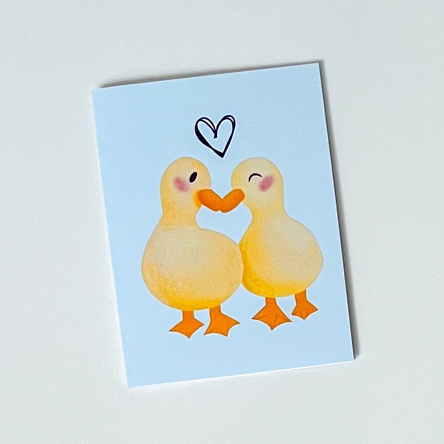 Card with two drawn ducks holding hands and a heart on a light blue background