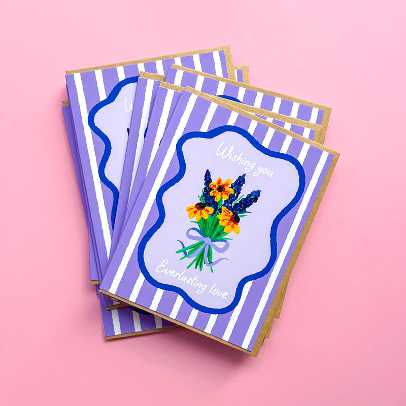 stack of wedding cards with hand drawn flowers and text 'wishing you everlasting love' on a purple striped background with Kraft envelope against pink background