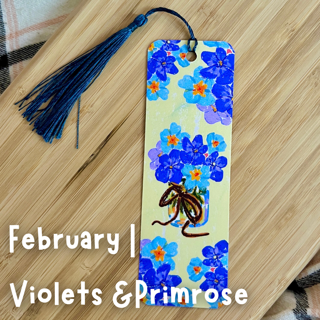 Floral bookmark with tassel on a wooden surface, featuring blue flowers and text 'February | Violets & Primrose'.