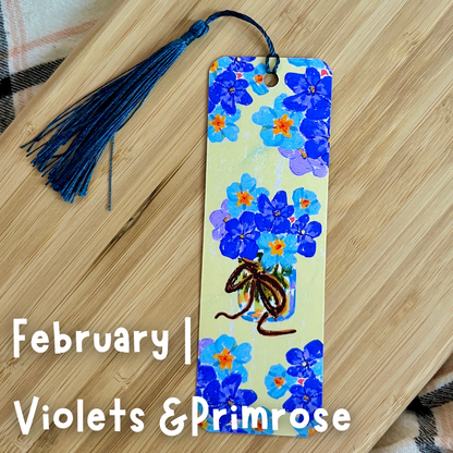 Floral bookmark with tassel on a wooden surface, featuring blue flowers and text 'February | Violets & Primrose'.