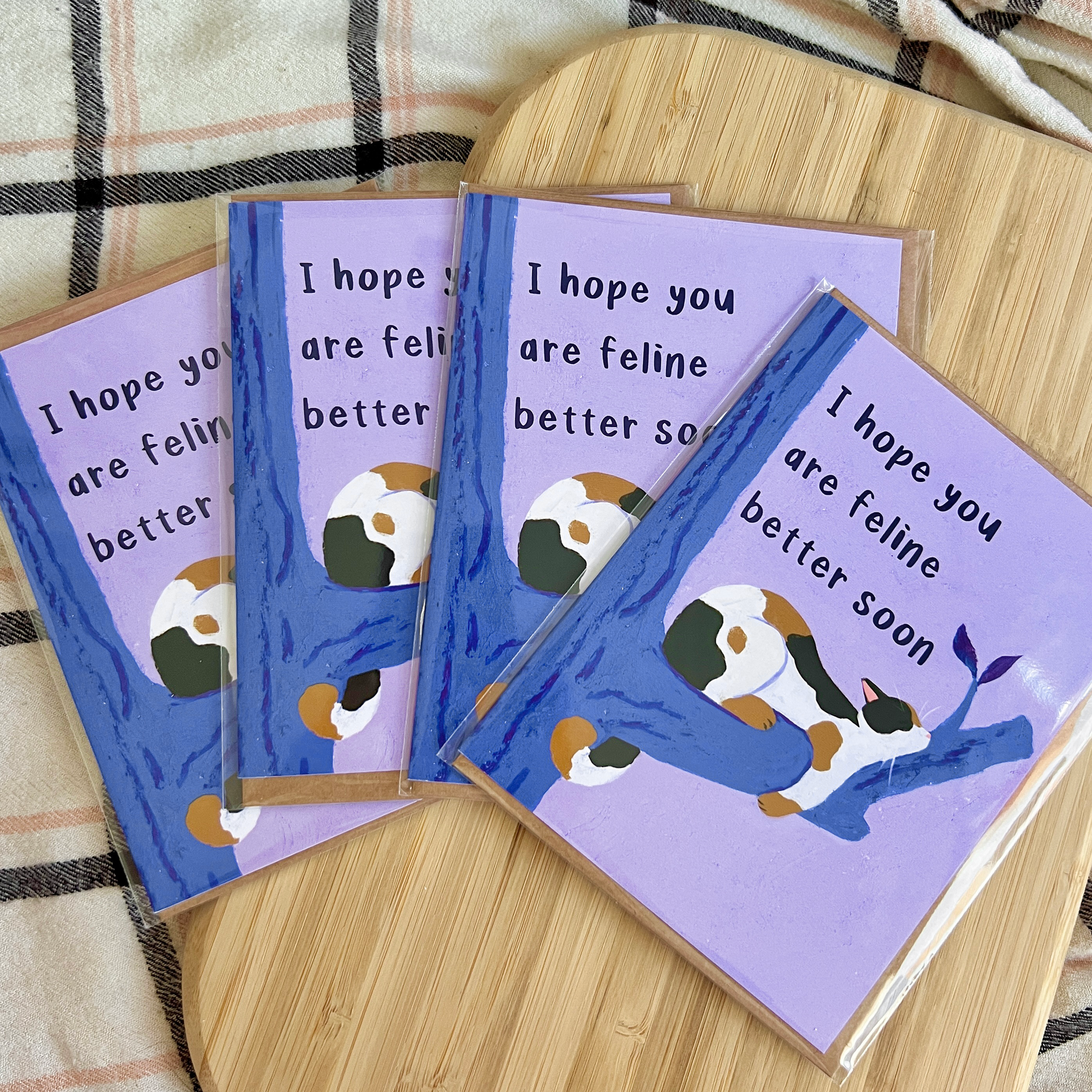 Four greeting cards with a cat illustration and text 'I hope you are feline better soon' on a wooden surface.