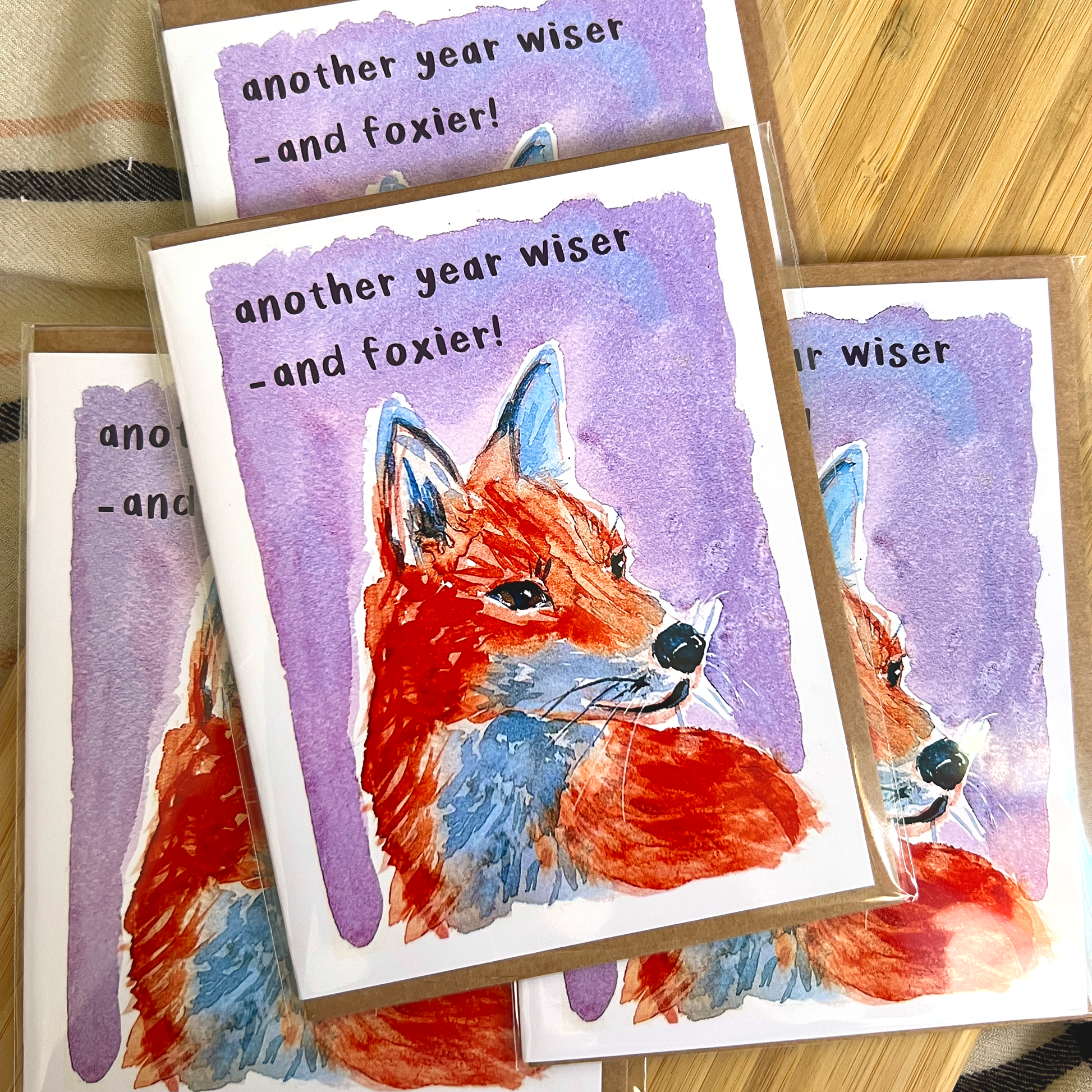 Greeting cards with fox illustration and text 'another year wiser and foxier!' on a wooden surface.