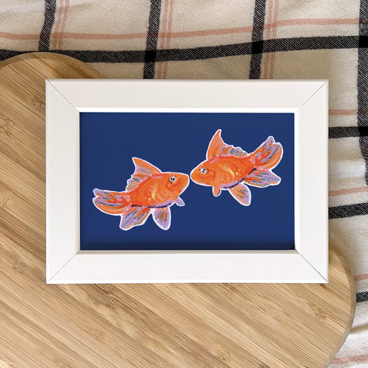 Framed artwork of two goldfish on a blue background, placed on a wooden surface with a plaid fabric background.