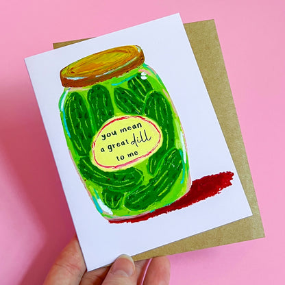 hand holding a greeting card with a pickle jar illustration and text 'You Mean A Great Dill To Me' on a pink background