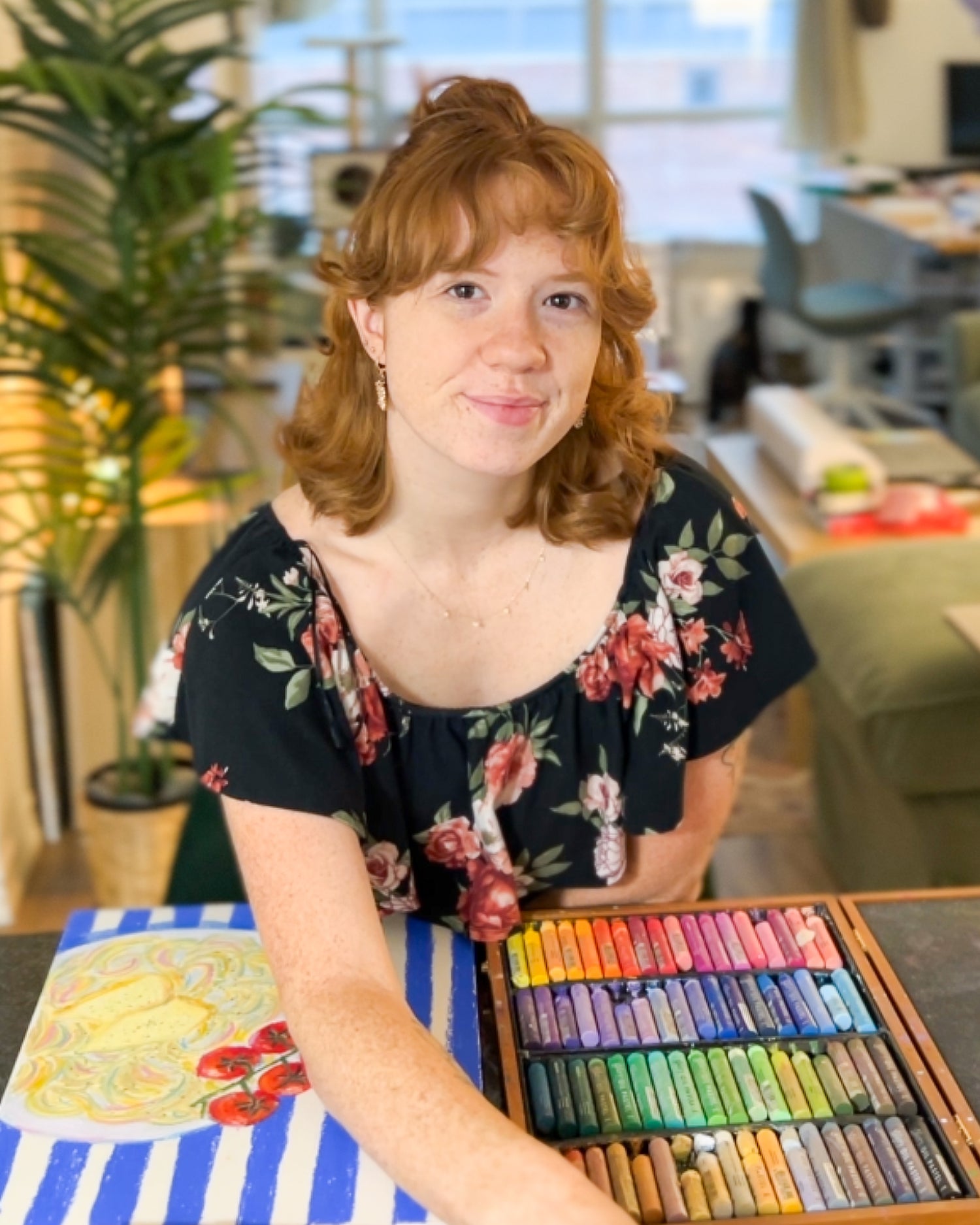 Person with red hair sitting at a table with pastel crayons and art supplies in an indoor setting.