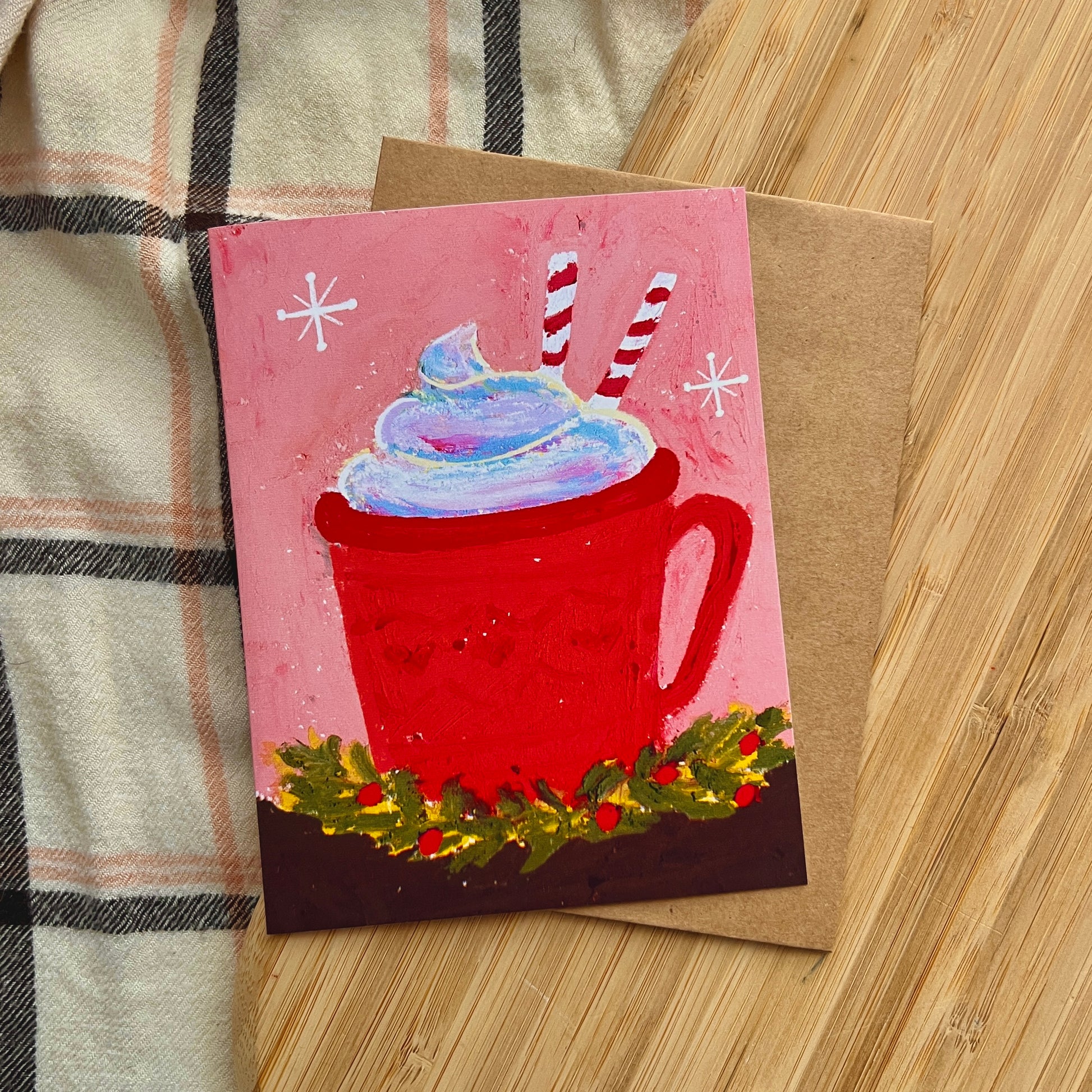 Card with a red mug of hot chocolate and whipped cream on a wooden surface.