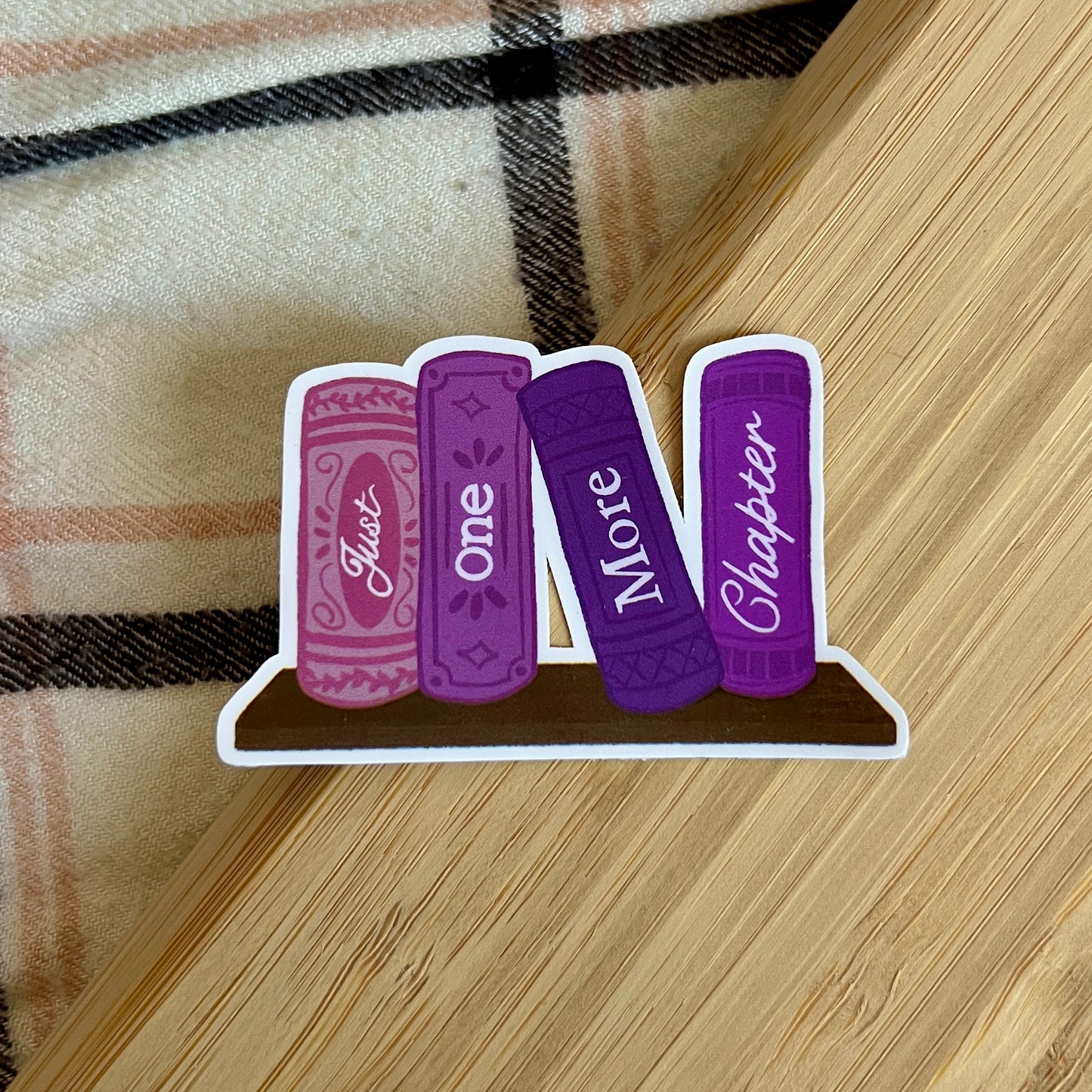 Sticker with purple books labeled 'Just One More Chapter' on a wooden surface.